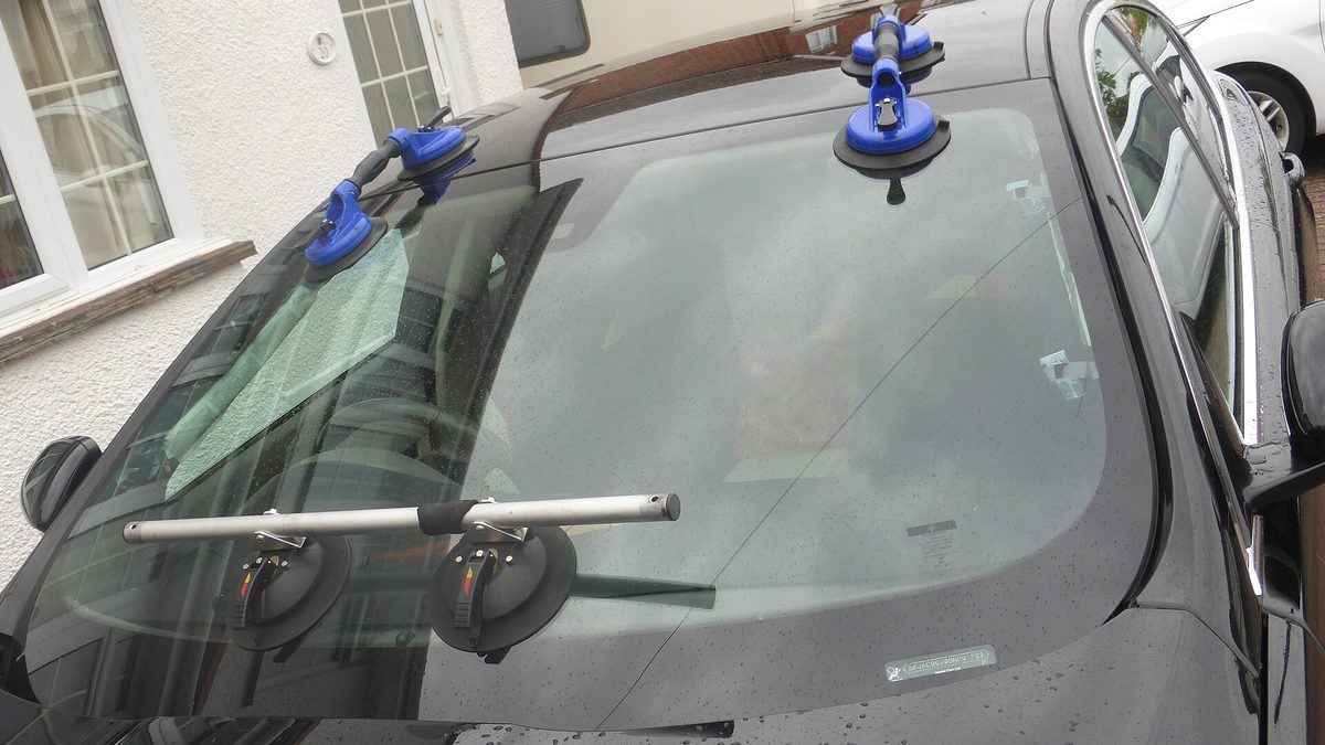 Neue Windschutzscheibe mit blauen Saughebern beim Einbau an einem schwarzen Auto