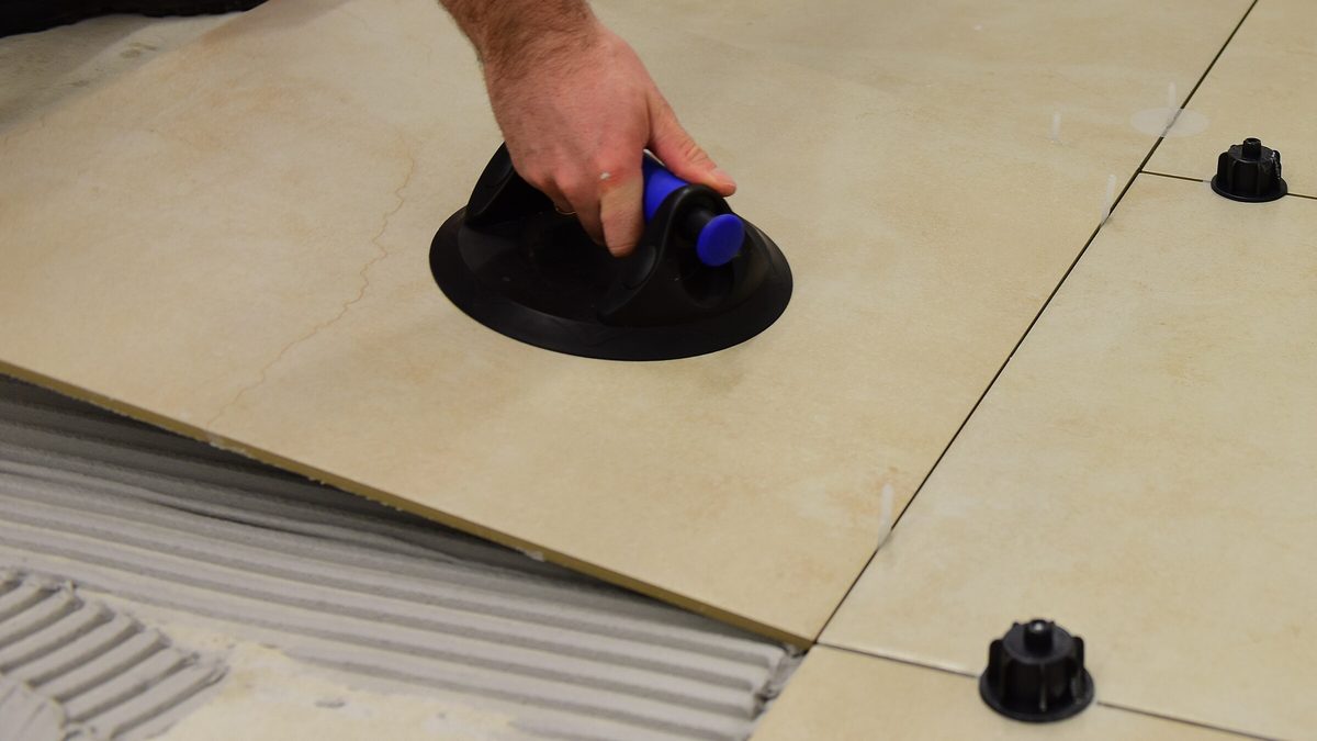 Hand setzt große beige Bodenfliese mit Saugheber, Nivellierclips und Fliesenkleber im Zahnbett