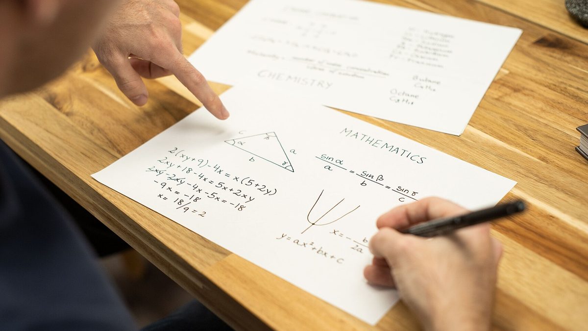 Lehrkraft zeigt auf ein Mathematik-Arbeitsblatt, während daneben jemand Formeln notiert