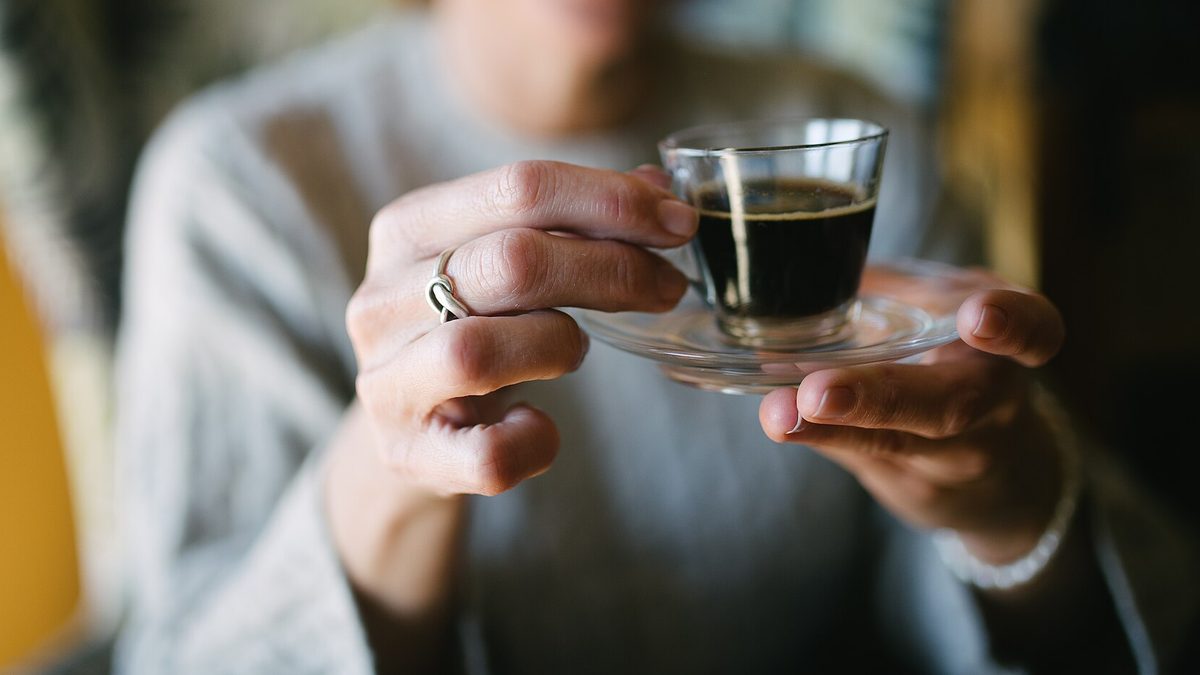 Hände reichen eine gläserne Tasse Espresso auf einer Untertasse entgegen