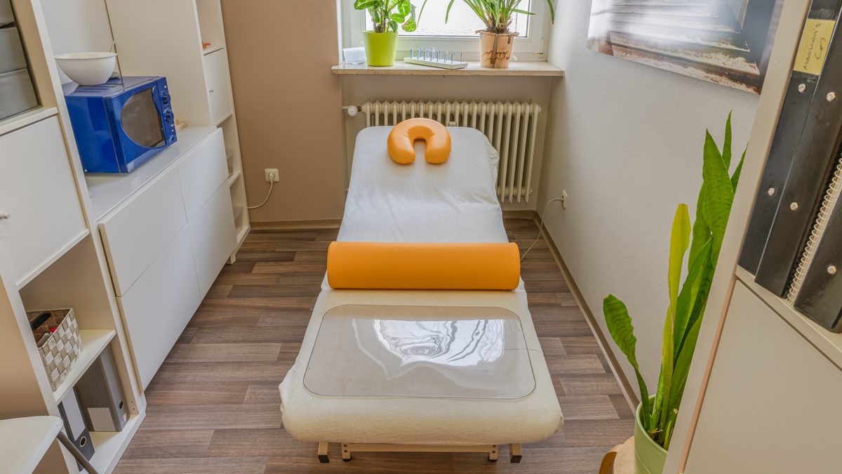 Heller Behandlungsraum einer Therapiepraxis mit Behandlungsliege, Lagerungsrolle und Grünpflanzen