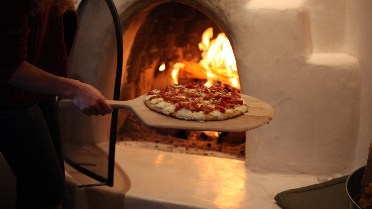 Pizza mit Tomaten wird auf einem Holzschieber in den brennenden Holzofen geschoben
