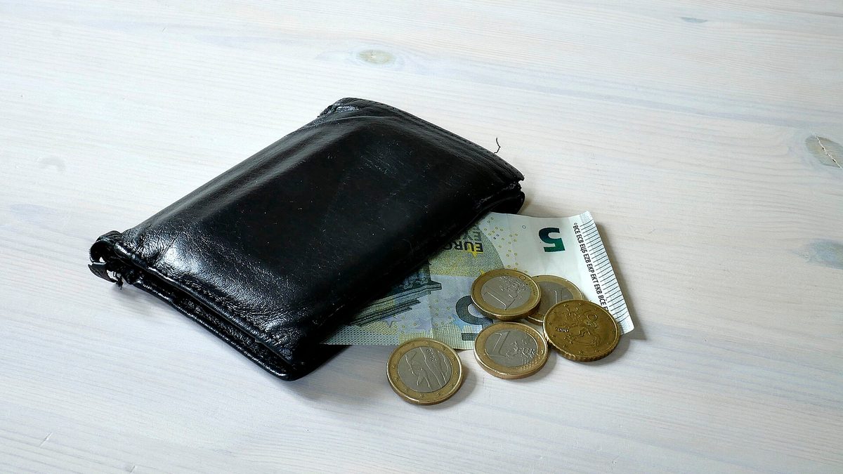 Schwarze Geldbörse mit einem Fünf-Euro-Schein und wenigen Euro-Münzen auf hellem Holztisch